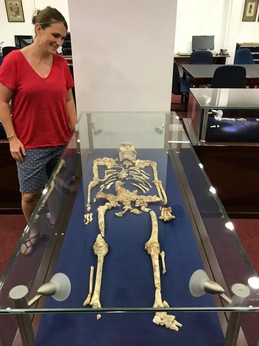 a woman in a red shirt stands next to a human-like skeleton in a glass case