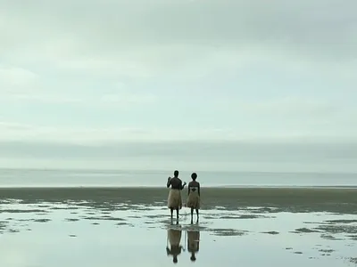Two people in grass skirts stand on the shore, facing the ocean under an overcast sky.