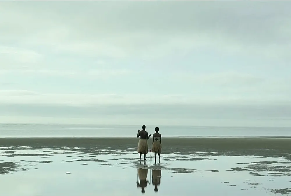 Two people in grass skirts stand on the shore, facing the ocean under an overcast sky.