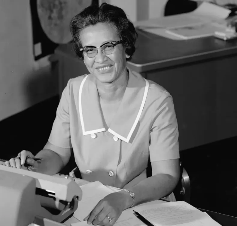 A black and white photo of Katherine Johnson sitting at a desk in front of a typewriter and open notebook.
