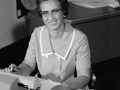 A black and white photo of Katherine Johnson sitting at a desk in front of a typewriter and open notebook.