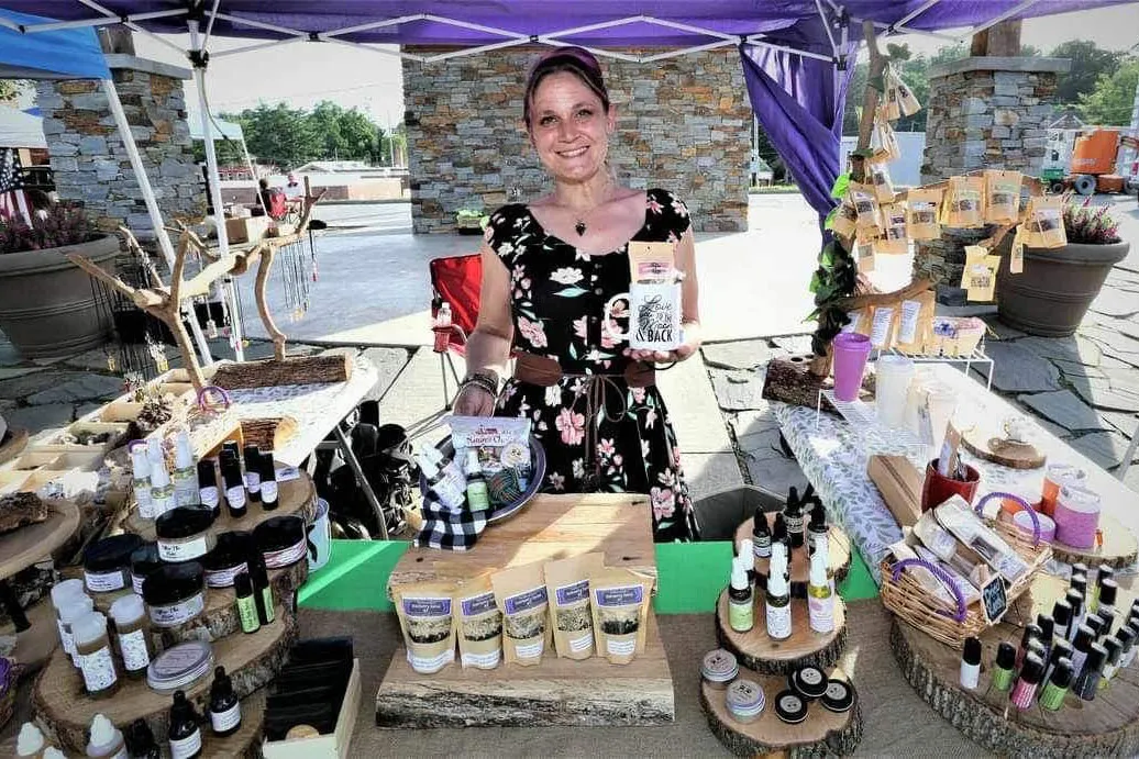 A woman smiles, holding up products in an outdoor market book. Her table displays include small glass containers and brown paper bags.
