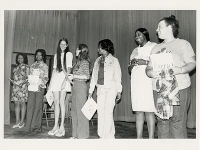 Candid photo of seven women holding papers standing in a line on stage.