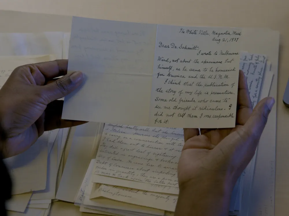 Two hands holding a card with handwritten information. A stack of other handwritten papers is visible on the table below.