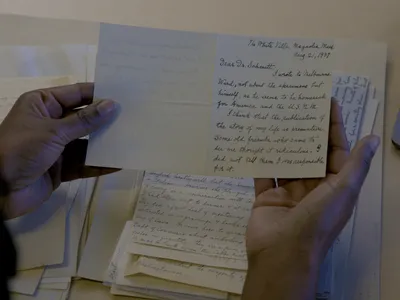 Two hands holding a card with handwritten information. A stack of other handwritten papers is visible on the table below.